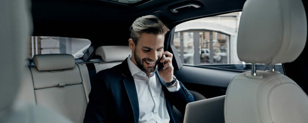 Discussing business details. Handsome young man in full suit talking on smart phone and smiling while sitting in the car