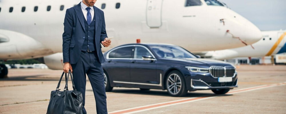 Stylish man in a business suit looking at his phone while standing in front of a car near a plane